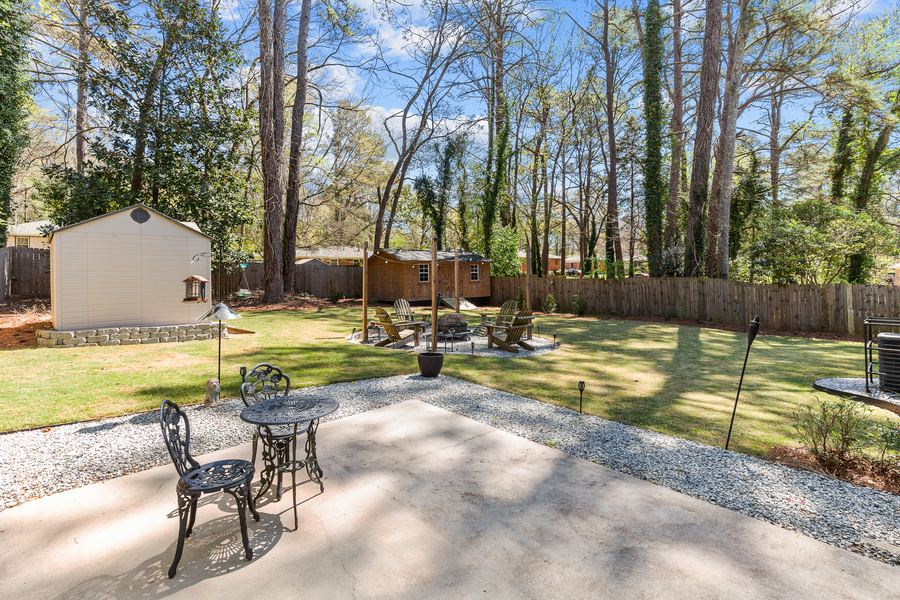 A shaded backyard patio with a bistro table, firepit seating and mature trees